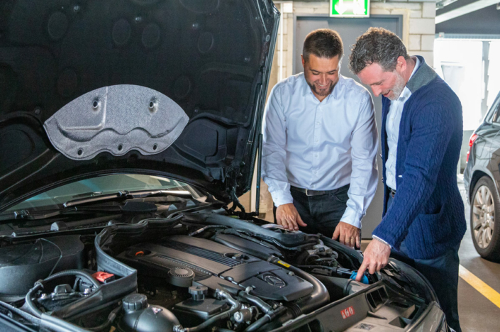 Two men are looking at the hood of a car.