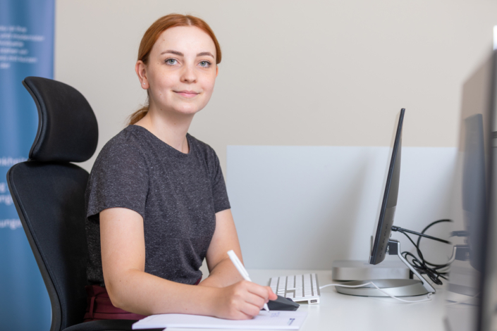 A woman sits at a desk with a computer and a pen.